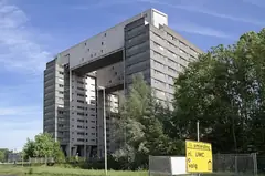Голяма сива сграда на студентско общежитие с мостова надстройка, дървета, ограда и жълт пътен знак отпред