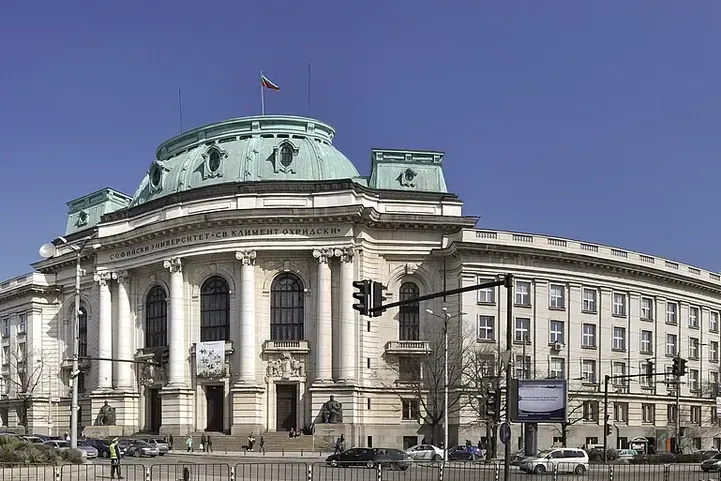 Панорама на сградата на Софийския университет „Св. Климент Охридски“ със зелен купол, български флаг и улица отпред