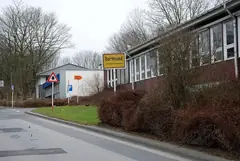 Табела „Dortmund, Ortsteil Eichlinghofen“ пред ниски сгради с много прозорци, улица, храсти и голи дървета