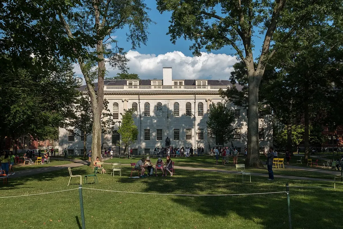 Бяла каменна сграда University Hall в Харвард Ярд с поляна, дървета, цветни столове и посетители през лятото